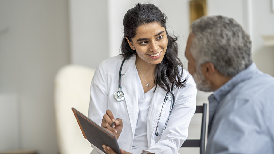 Photograph of a doctor in a lab coat, holding an ipad and speaking with a patient.